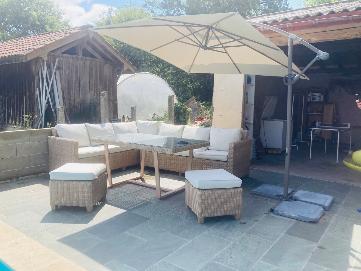 An outdoor seating area features a modern wicker sectional sofa with plush cushions, surrounding a glass-top table. An adjustable umbrella provides shade, while a stone patio floor complements the setting. Various garden structures and greenery are visible in the background.