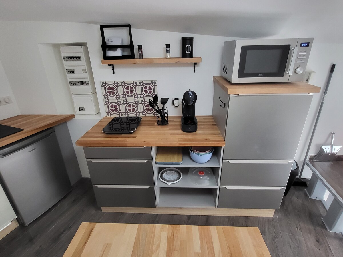 A compact kitchen area features modern appliances, including a microwave and coffee machine. A wooden countertop extends over cabinets with a mix of open and closed storage. Essential kitchen utensils and a decorative tile backsplash enhance the functional design.