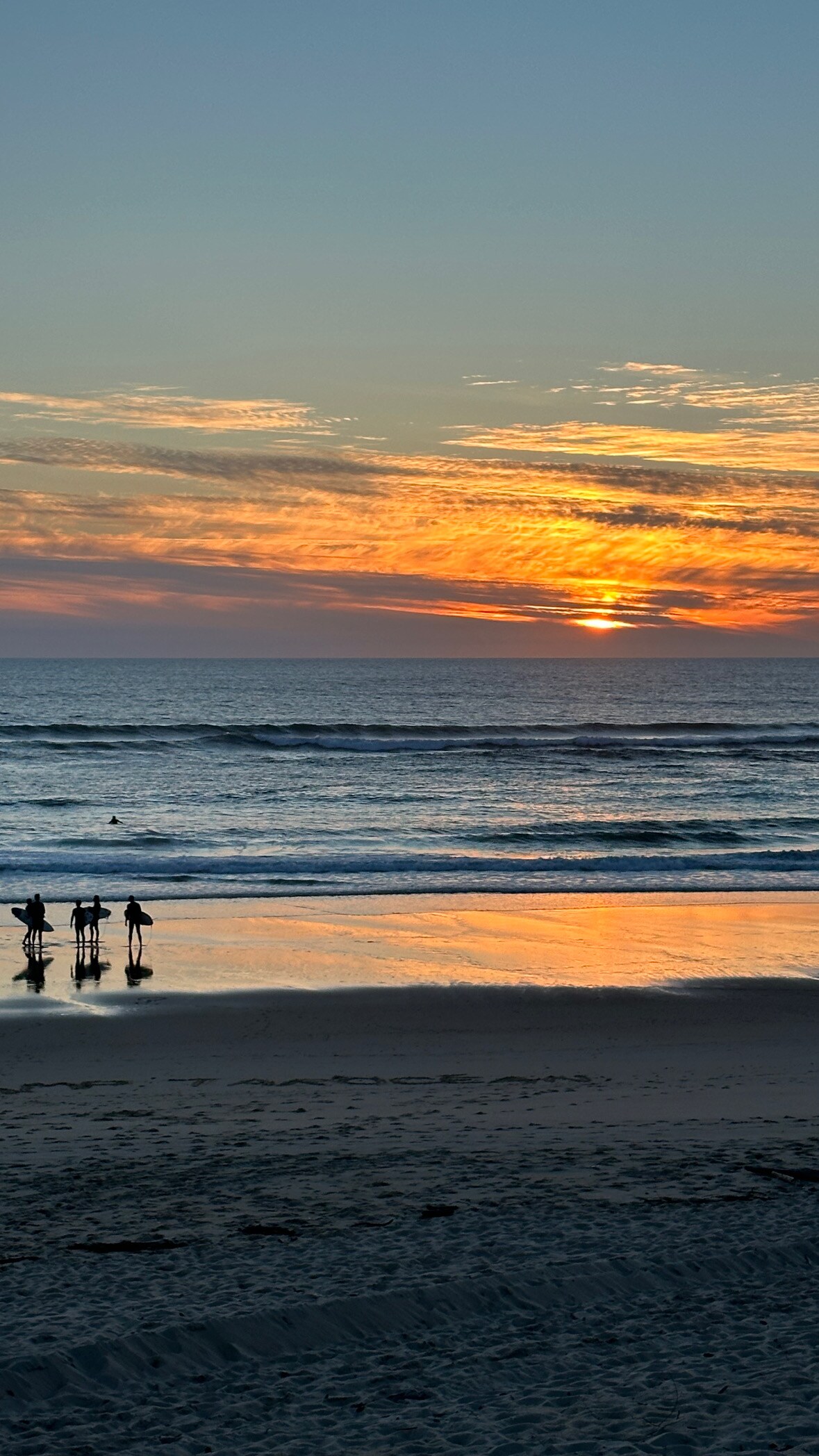 The image captures a beach scene at sunset, featuring silhouettes of individuals standing on the shore. The vibrant colors of the sunset reflect on the water, creating a serene atmosphere as gentle waves lap at the sandy beach.