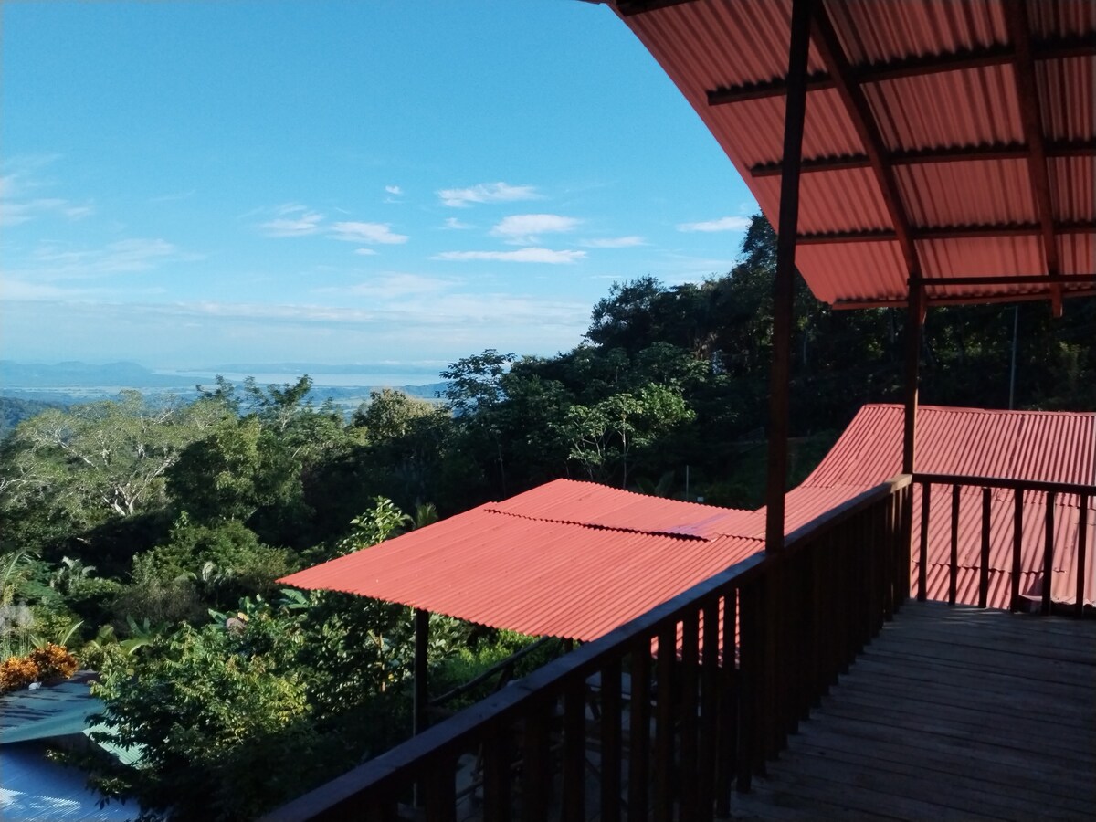 A wooden balcony offers a view of the surrounding mountains and lush greenery. Red metal roofs of nearby structures can be seen below, while the sky above is clear with a few scattered clouds. The distant coastline is visible, enhancing the natural environment.