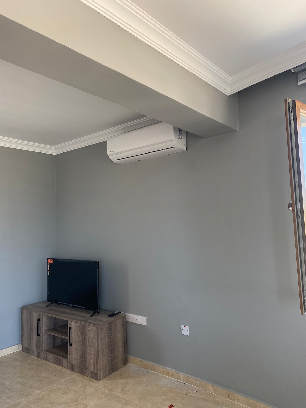 A simple living area is shown, featuring a mounted TV on a wooden stand. The walls are painted in a subtle gray, and natural light enters through a window on the right. An air conditioning unit is installed near the ceiling.