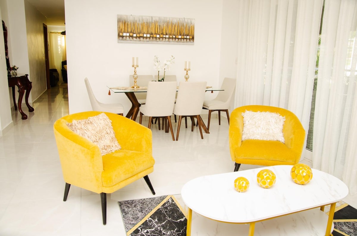 A bright living space features two yellow armchairs positioned beside a marble coffee table. A dining area with a large table and light-colored chairs is visible in the background, complemented by soft natural light filtering through sheer curtains.