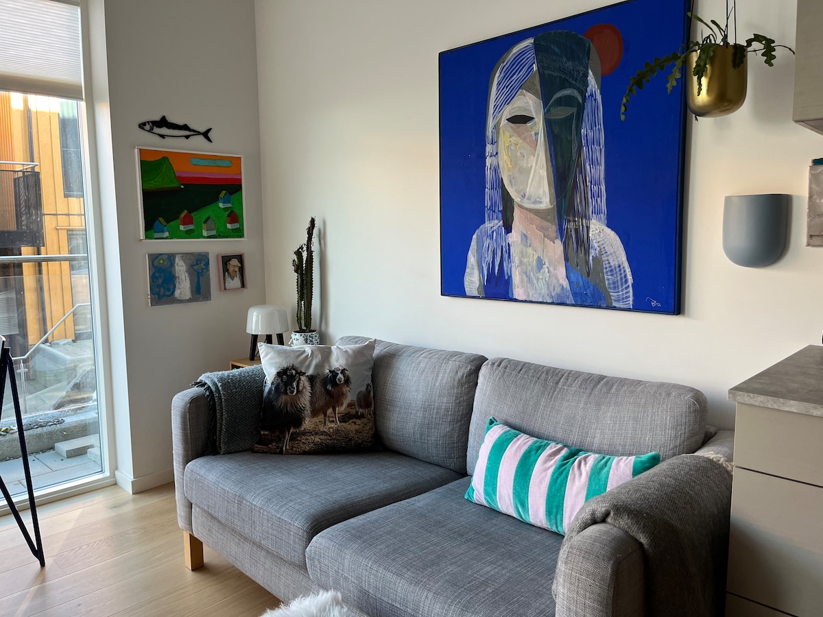 A living area features a grey corner sofa adorned with a striped cushion and decorative throw. A large artwork of a woman hangs on the wall, complemented by smaller artworks. Natural light is captured through floor-to-ceiling windows and a potted plant adds a touch of greenery.