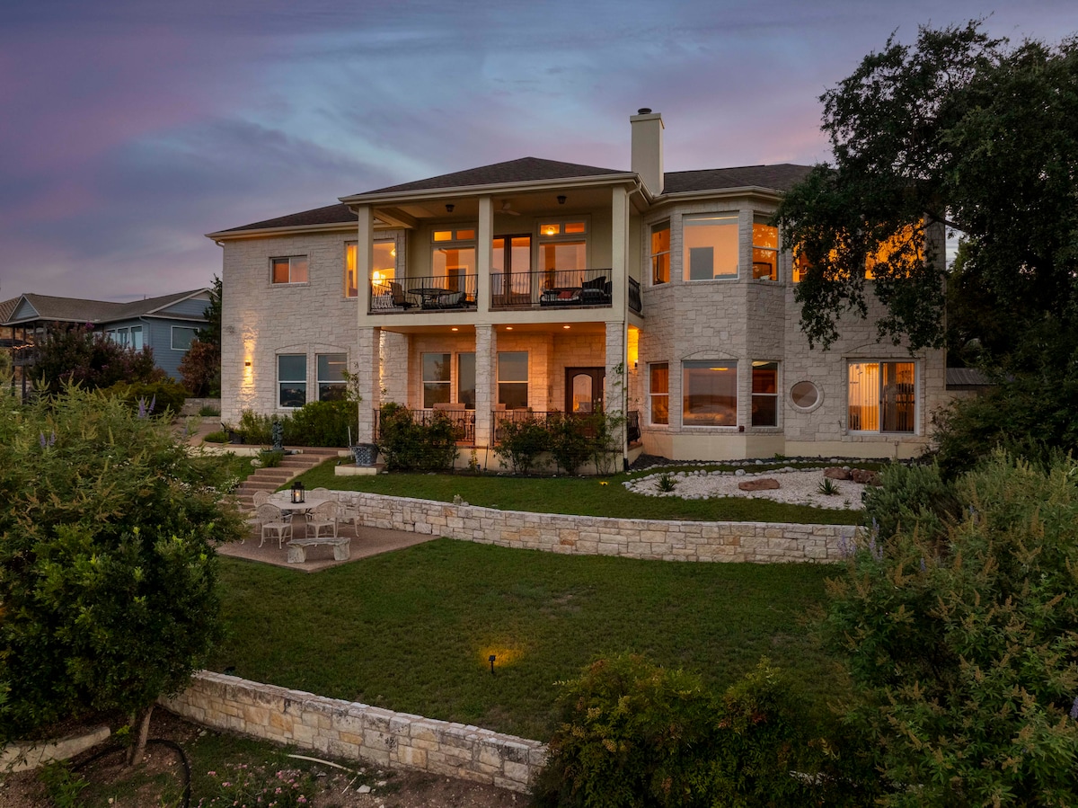 The exterior of The Sun Rise Lakehouse showcases a spacious two-story structure with a light stone facade. The balcony features seating arrangements, while soft orange light emanates from the windows, hinting at the inviting interior. Lush green landscaping frames the home, with a view of Lake Travis in the background.