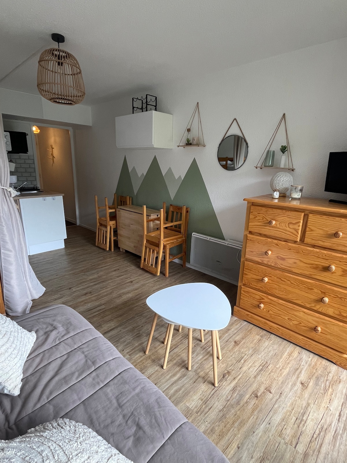 A studio living area features a seating space with a grey sofa and a light wood dresser. A small white table is positioned in front of the sofa, accompanied by a dining table with six wooden chairs. Green mountain-themed wall art is displayed alongside mirrors.