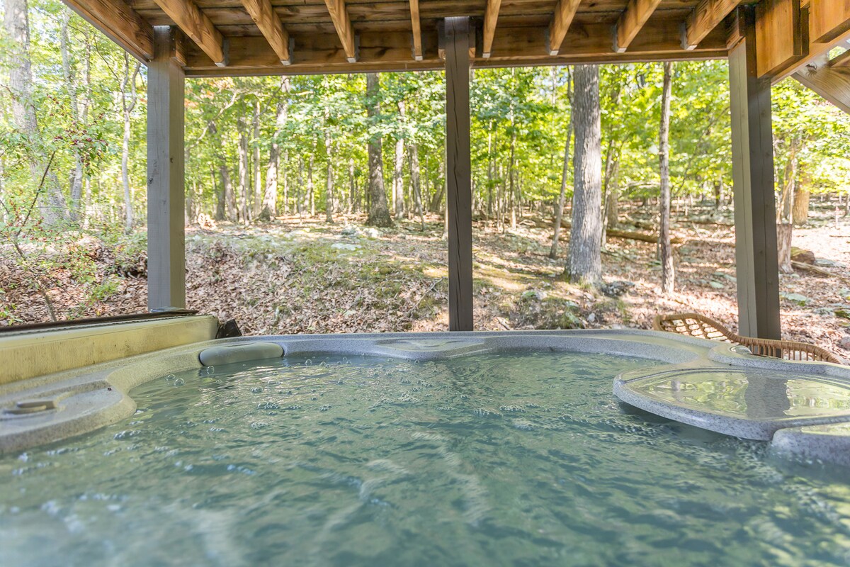 A hot tub is situated under a wooden structure, surrounded by lush green trees and natural foliage. Sunlight filters through the leaves, creating a serene atmosphere and inviting relaxation in the peaceful outdoor setting.
