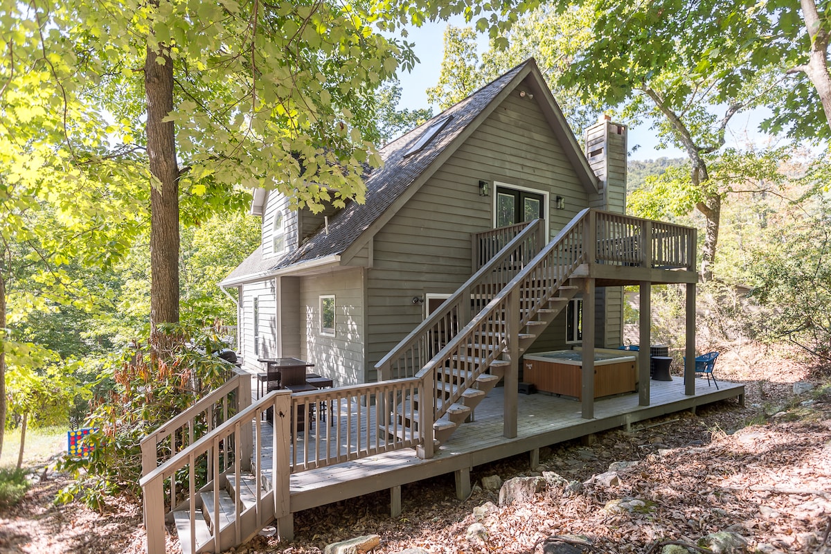 The exterior of the mountain retreat showcases a well-maintained two-story structure with a wooden deck. Steps lead up to the entrance, surrounded by lush greenery and natural foliage, creating a peaceful setting. The hot tub is visible on the deck, providing a relaxing outdoor option.