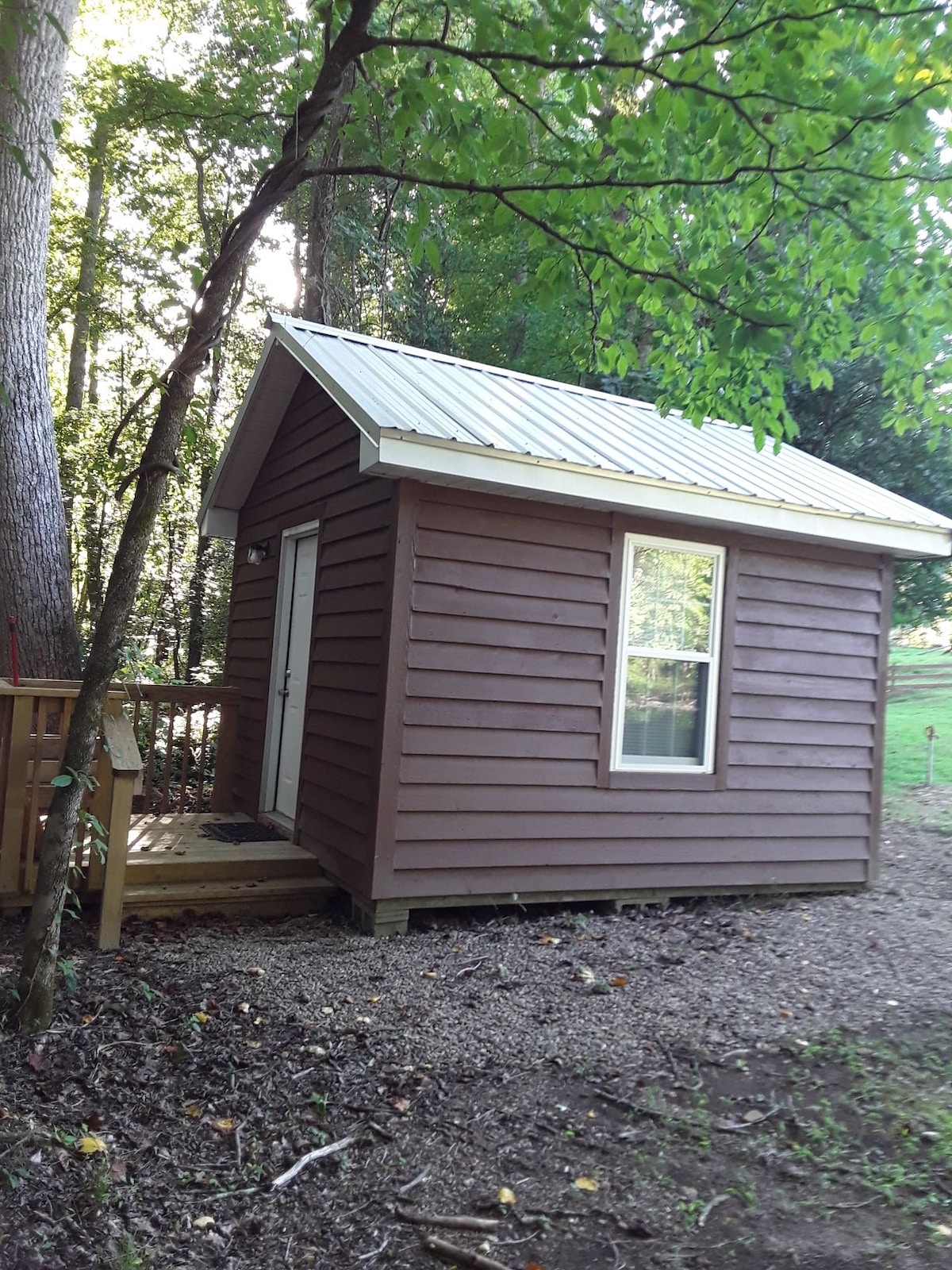 A small cabin with a brown wooden exterior is surrounded by trees. The entrance is accessible via a wooden deck with steps leading up. A single window allows natural light to enter the space, and the surrounding area is covered with gravel.