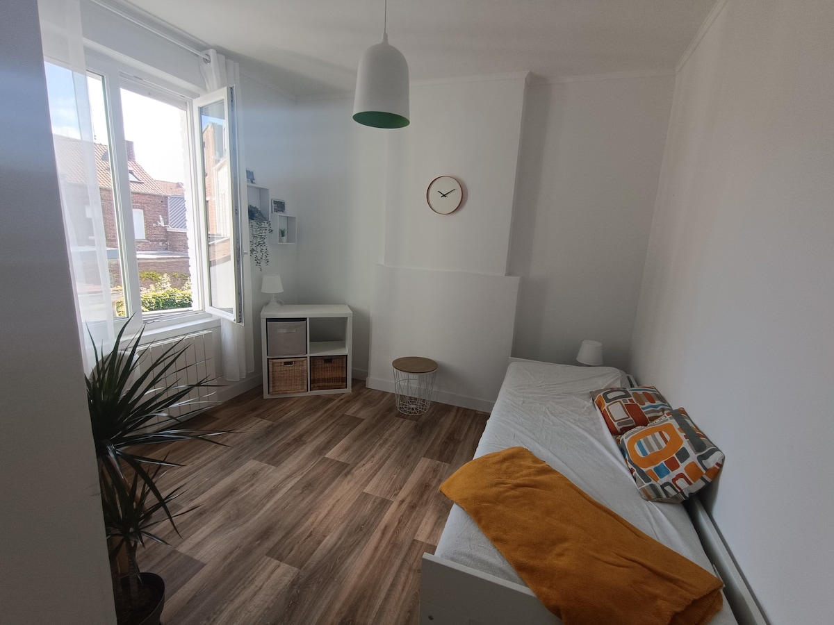 A bright room features large windows allowing natural light to fill the space. A single bed is positioned against the wall, complemented by a decorative orange throw. A small round table and a storage unit with cubes add functionality, while a potted plant brings a touch of greenery.