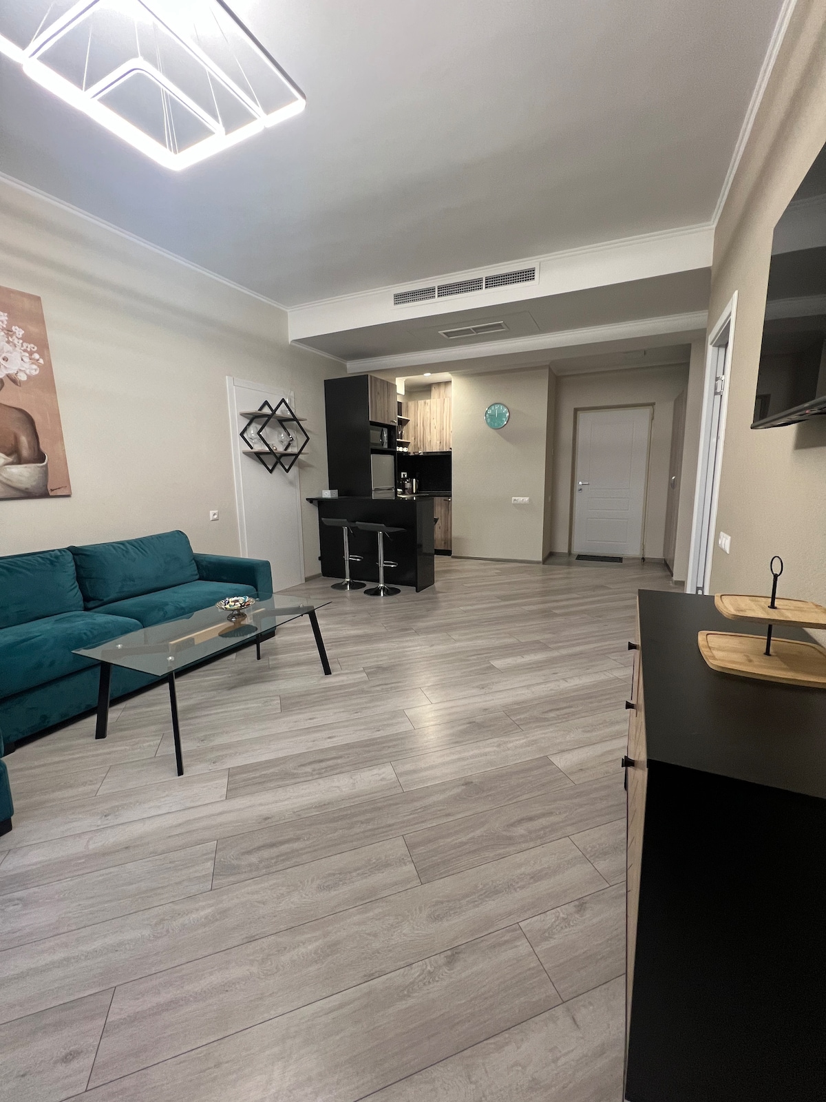A modern living space features a teal sofa and a glass coffee table. A kitchenette with bar stools is visible in the background, along with contemporary cabinetry. Natural light fills the room, highlighting the sleek flooring and minimal decor.