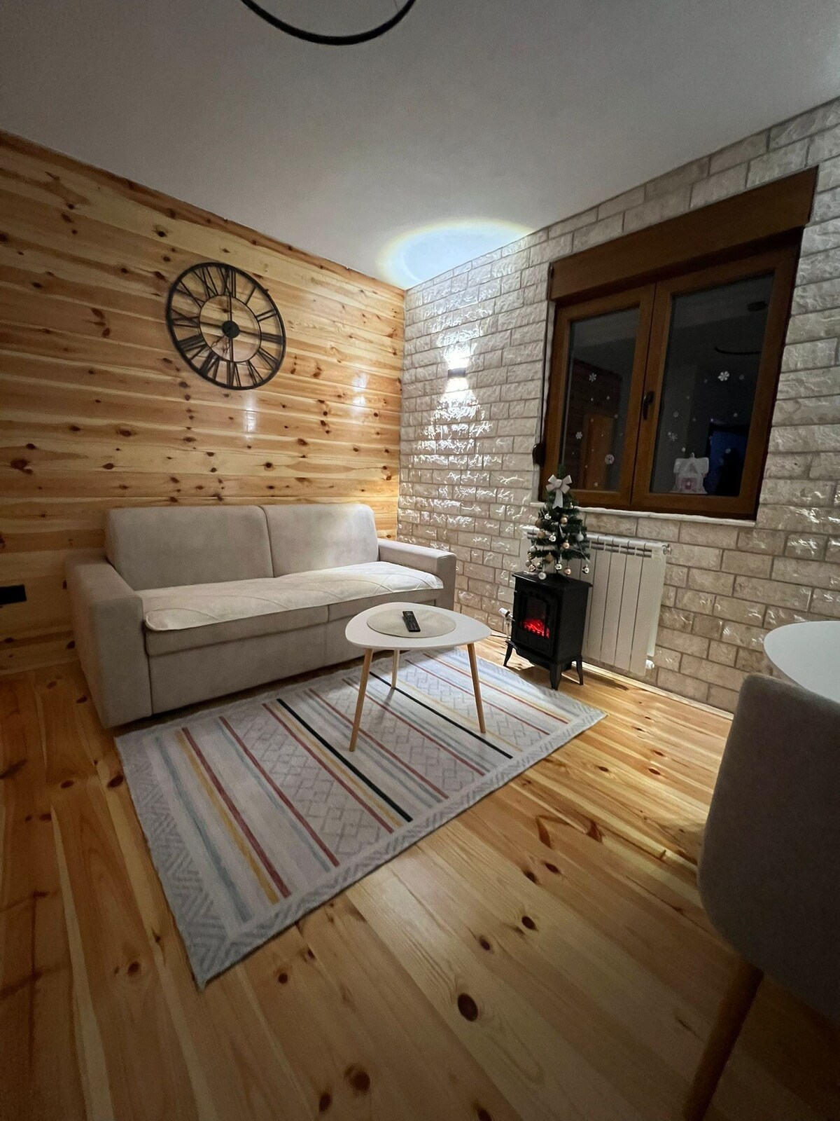 A cozy living area features a light-colored sofa and a small round table on a patterned rug. Soft lighting enhances the natural wood walls, while a decorative clock hangs above. A compact heater sits beside the window, which offers views of the outside.