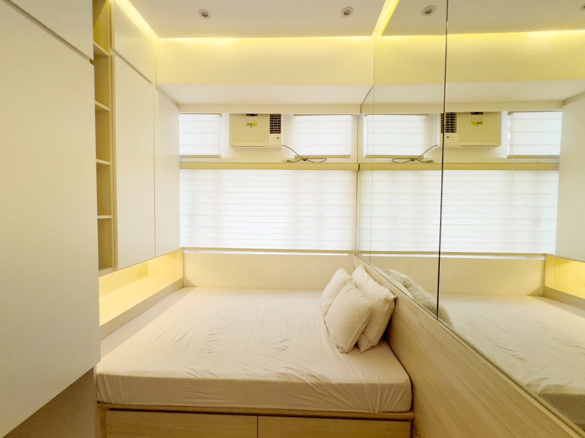 A bright bedroom features a spacious bed with a neutral-colored bedspread. Large mirrors create an illusion of openness. Two air conditioning units are visible above the window, which is covered by sheer white blinds, allowing natural light to filter through.