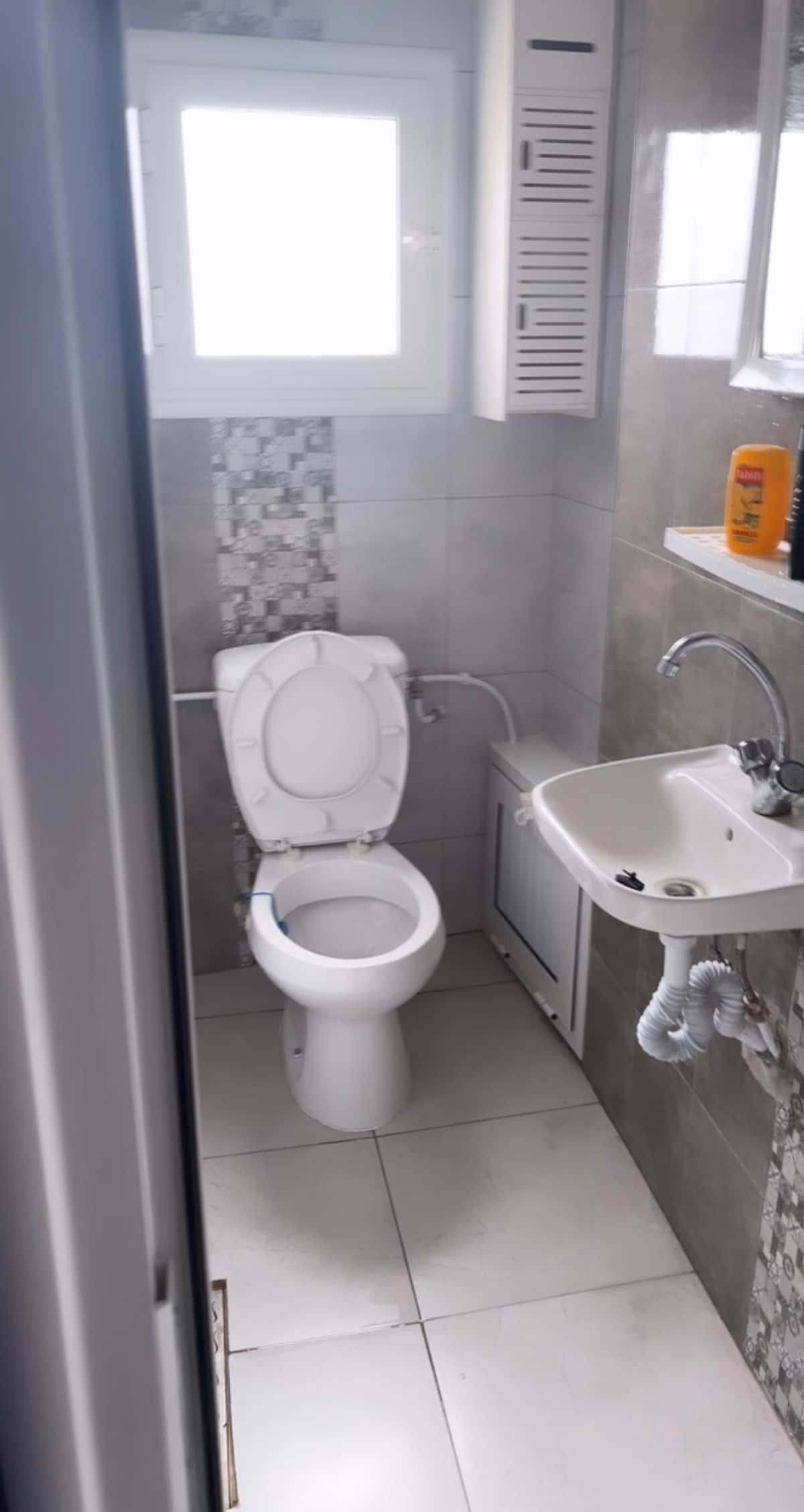 The bathroom features a standard toilet and a small sink with modern fixtures. Light reflects off the tiled walls, which include a decorative mosaic panel. A window allows natural light to enter, enhancing the space's overall brightness.