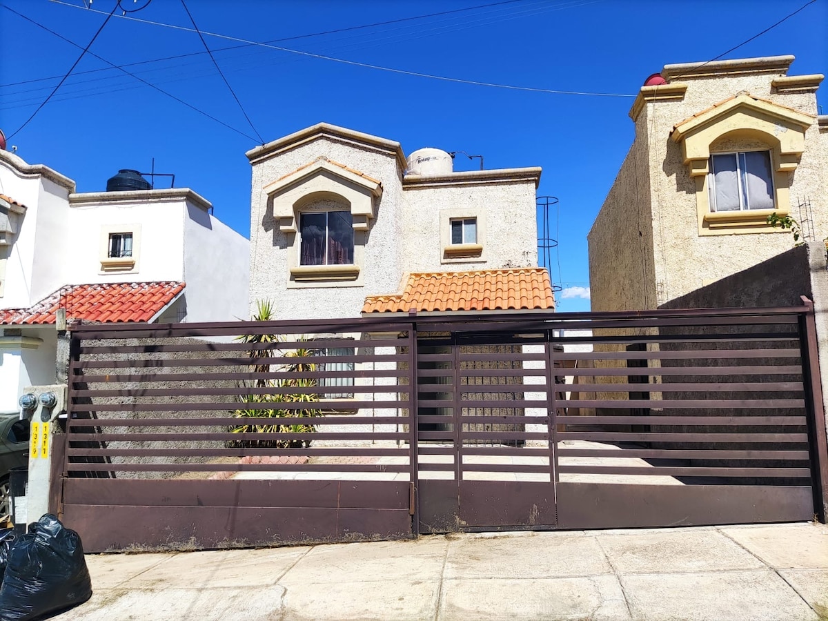 The exterior of a welcoming home is shown, featuring a gated entrance with horizontal slats. The façade includes details like a tiled roof and arched windows. Surrounding greenery adds to the visual appeal, and blue skies fill the background.