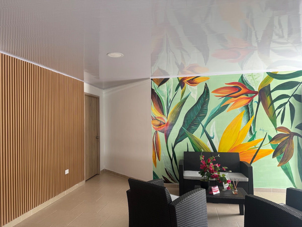 A vibrant living space features a large mural of tropical flowers on one wall, with a sleek wooden panel accent on the opposite side. A dark wicker seating area is arranged with a small table, complemented by a decorative arrangement of flowers.