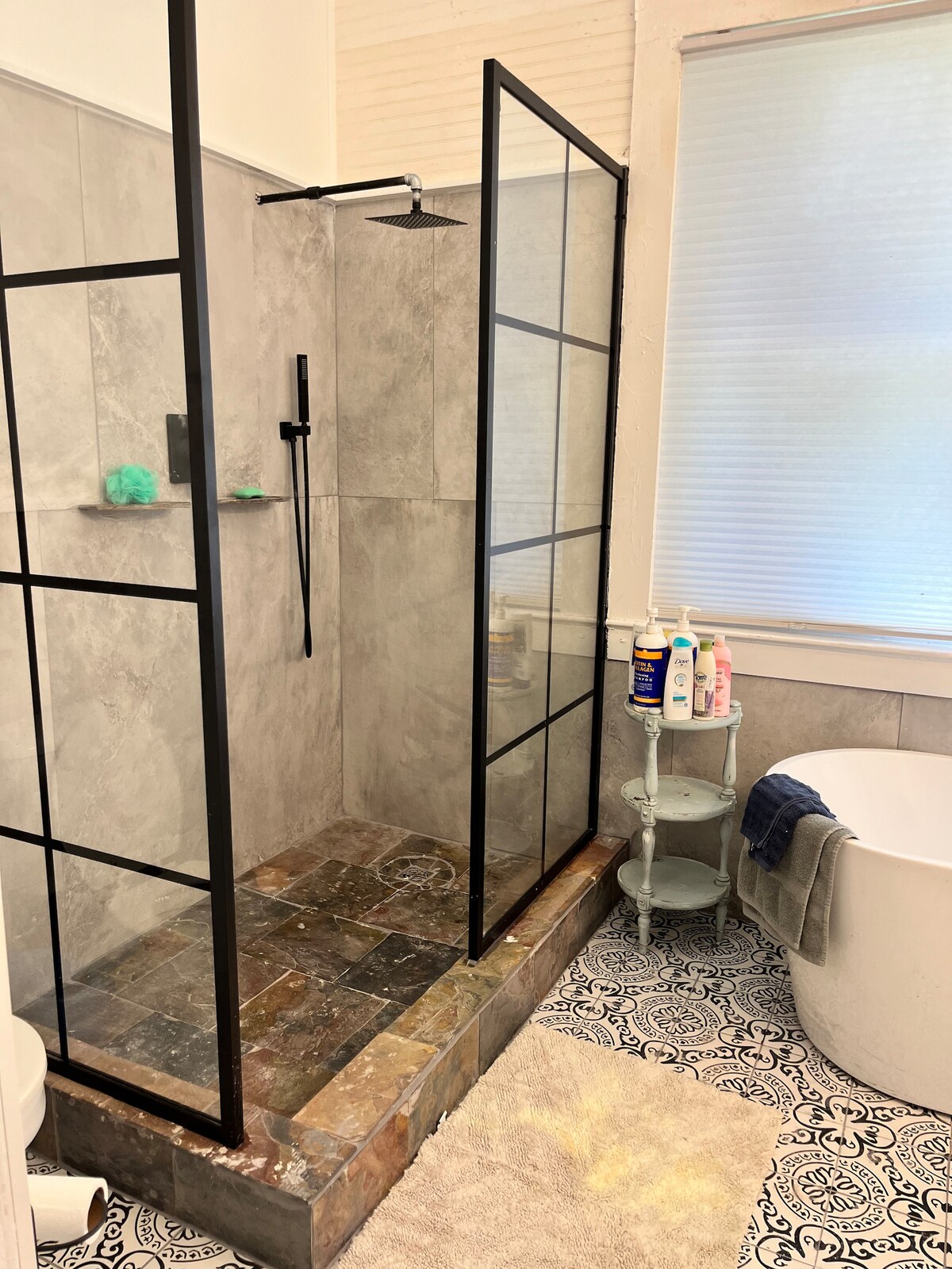 A modern bathroom features a glass-enclosed shower with a rainfall showerhead and a freestanding bathtub. Natural light filters through a window, illuminating the gray tile and patterned floor. A small cart holds various toiletries, enhancing functionality within the space.