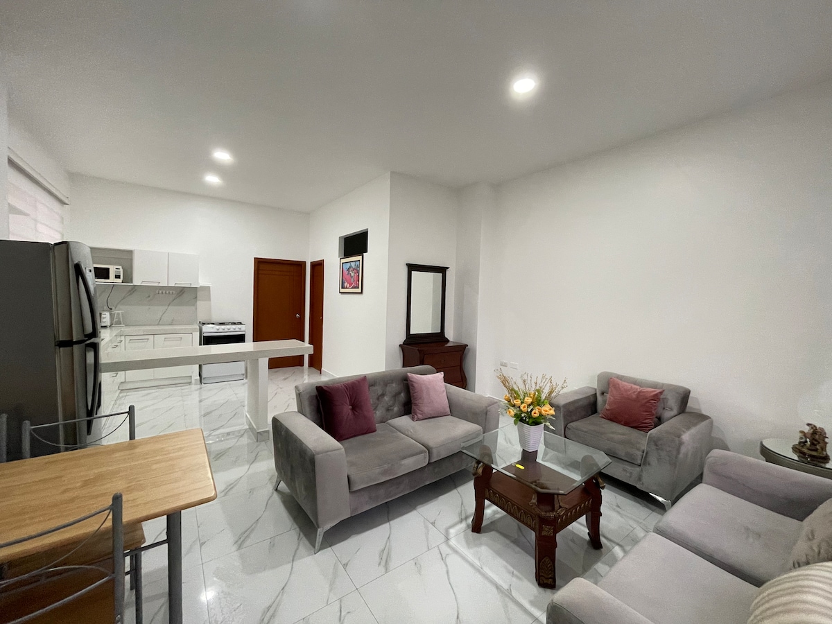 A bright living area features two comfortable sofas adorned with decorative pillows and a wooden table. A simple dining set is positioned nearby. The kitchen is visible in the background, showcasing modern appliances and a clean, white aesthetic. Light is softly illuminated throughout the space.
