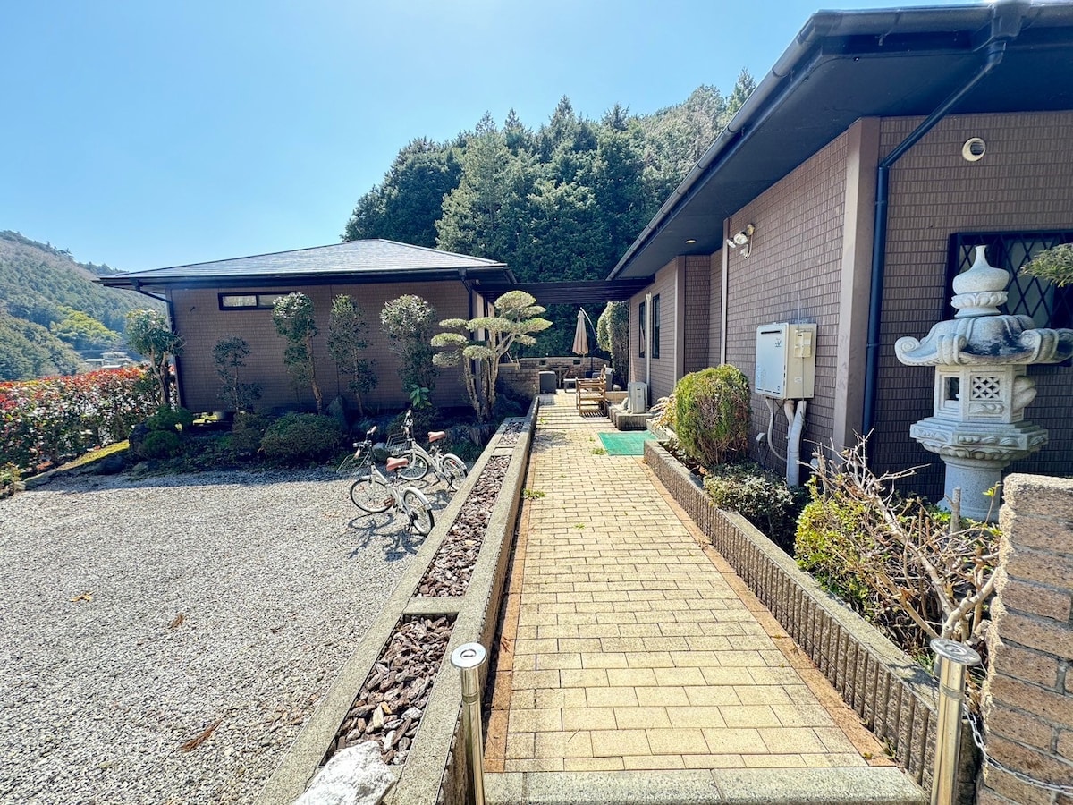 A pathway lined with bricks leads to a spacious outdoor area surrounded by greenery. Two buildings are visible, with one featuring a patio umbrella and a stone lantern nearby. Bicycles are placed on the gravel area, enhancing the tranquil ambiance.