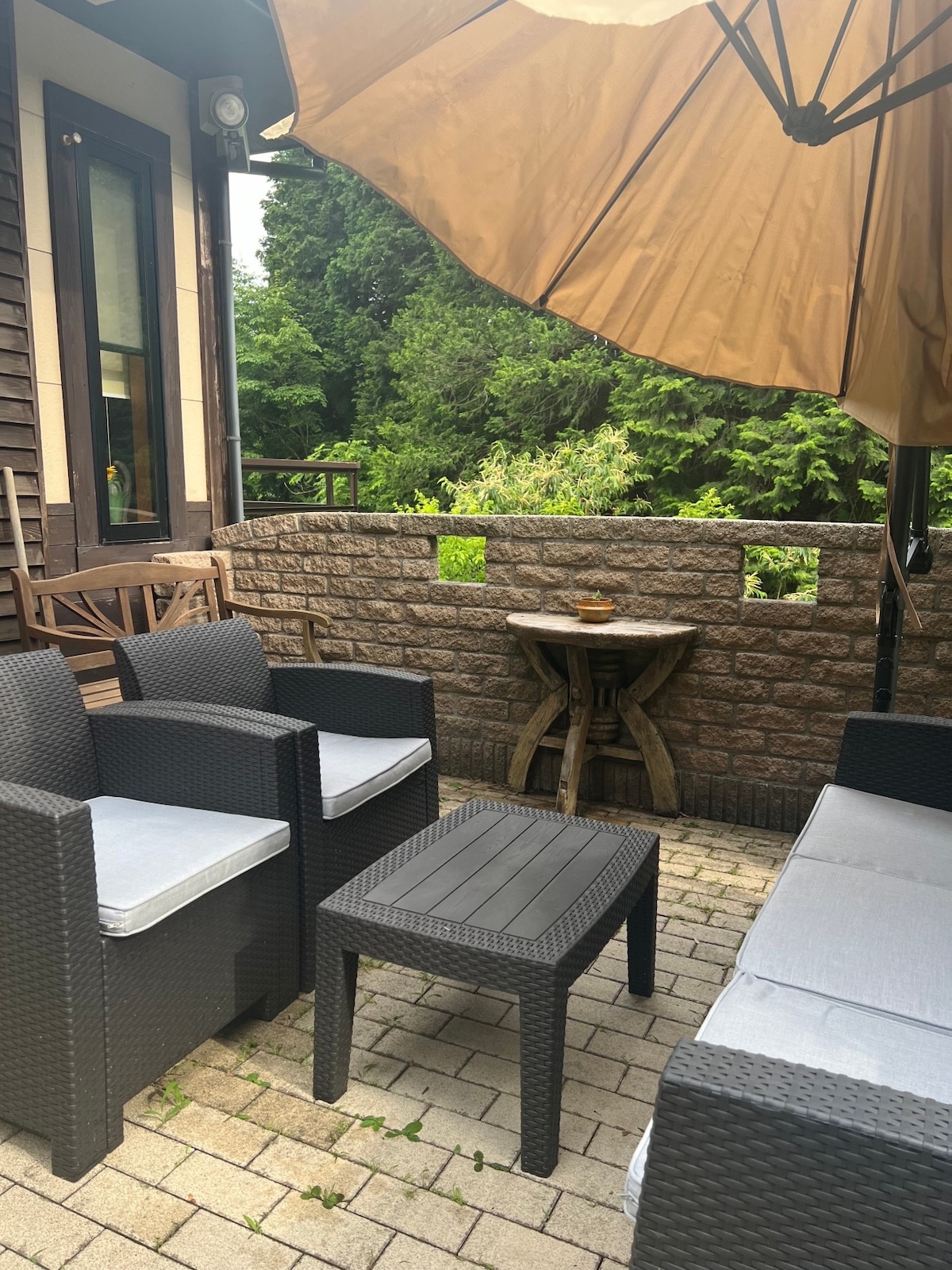 An outdoor seating area is presented with four chairs and a low table on a paved surface. A large umbrella provides shade while greenery is visible in the background, creating a secluded feel. The stone wall adds a rustic touch to the space.