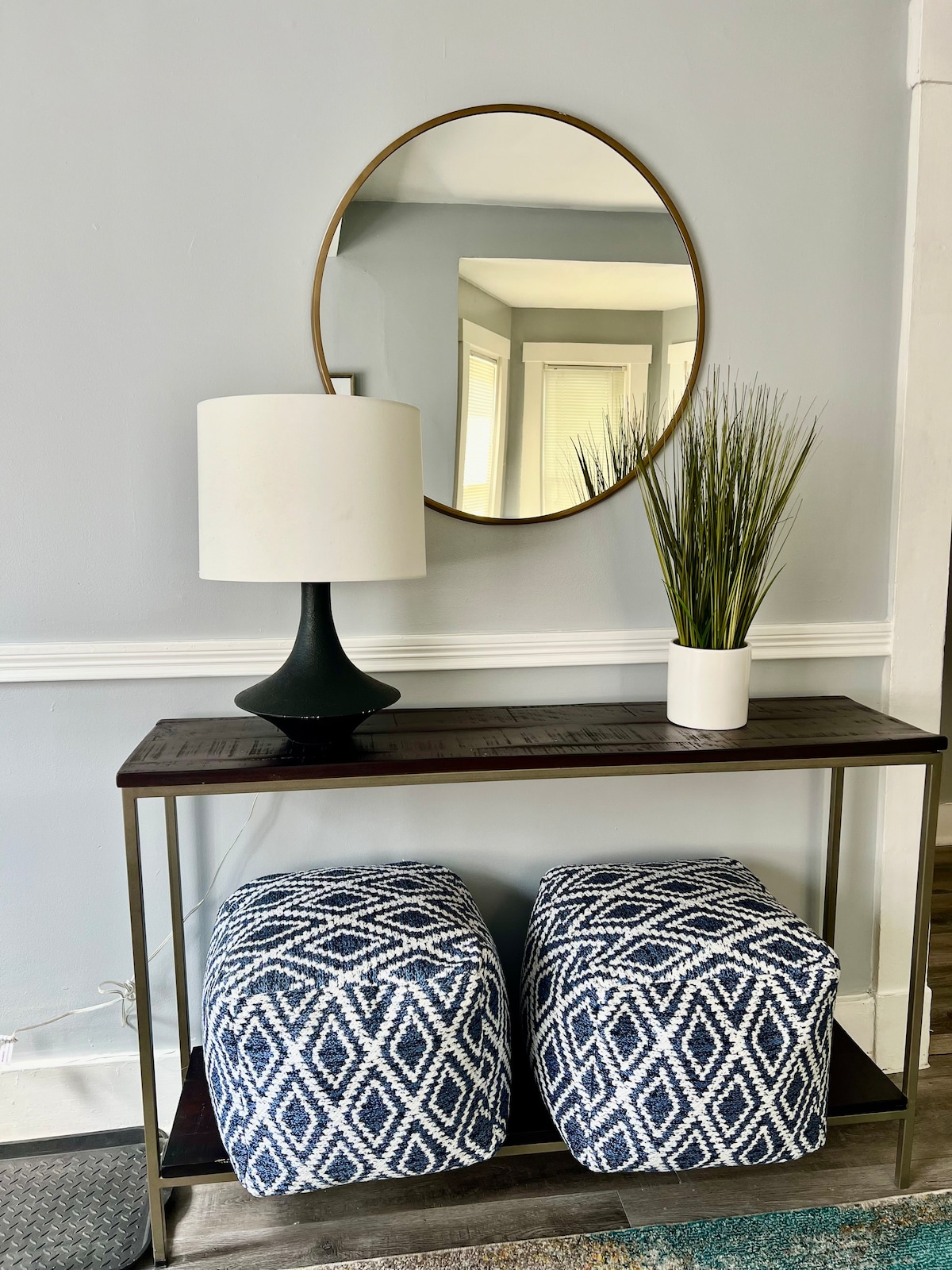 A modern console table is positioned against a light grey wall, featuring a circular mirror above. A black lamp provides ambient lighting, while two patterned poufs with geometric designs offer additional seating. A small white planter with greenery sits beside the lamp.
