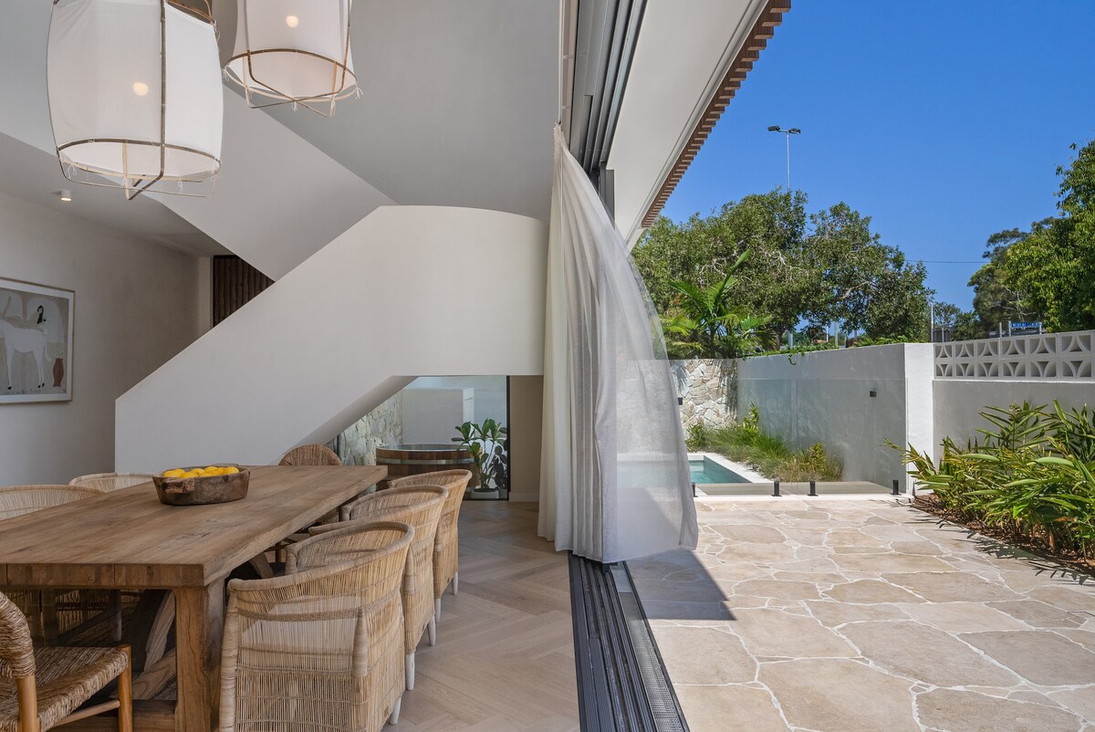 An indoor dining area features a large wooden table surrounded by natural wicker chairs. Floor-to-ceiling glass doors slide open to reveal a sunny outdoor space, where a pool is visible amidst lush greenery. A staircase elegantly descends in the background.