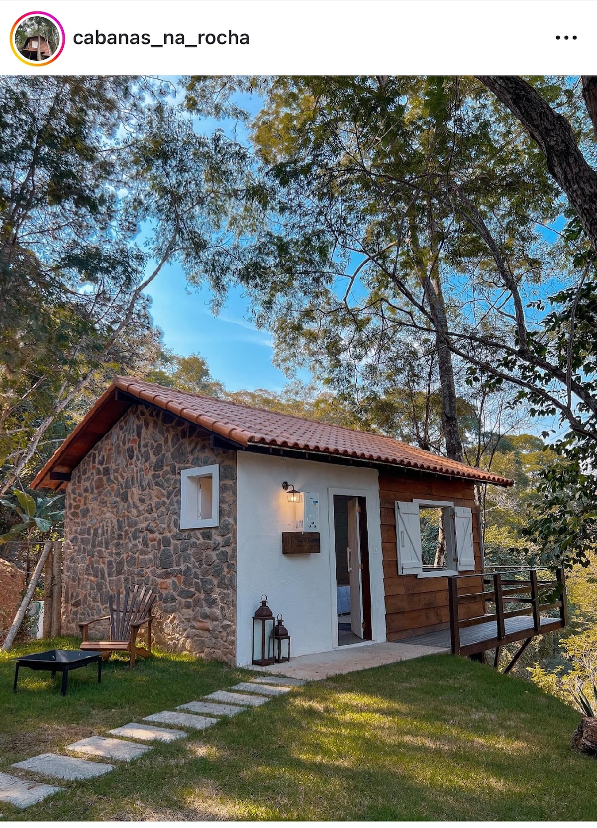 A small cabin features a blend of stone and wooden exteriors, set amidst tall trees. A pathway of stepping stones leads to a wooden porch, furnished with a chair and a lantern, surrounded by a grassy area.