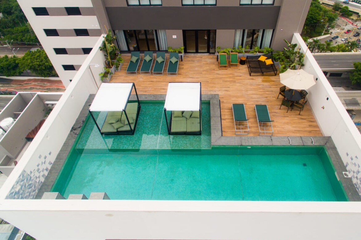 An aerial view of a rooftop pool area features a clear swimming pool surrounded by sun loungers and potted plants. Two transparent cabanas sit on the pool deck, while a dining table with chairs is positioned in one corner, offering space for relaxation.