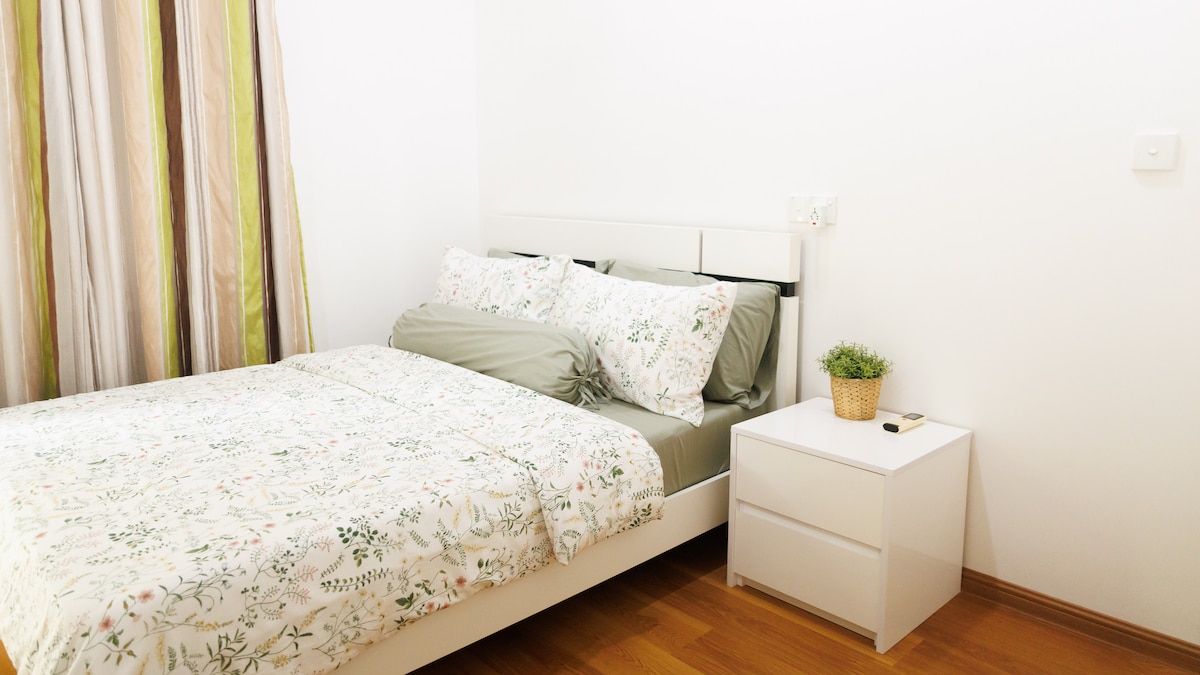 A well-appointed sleeping area features a spacious bed with light-colored floral bedding. A small nightstand positioned beside the bed includes a decorative plant. Natural light filters through sheer curtains, illuminating the warm wooden flooring and neutral wall color.