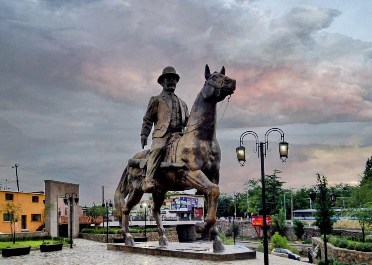 A large statue of a horseman on horseback stands prominently against a dramatic sky filled with clouds. The statue is set on a stone base, and street lamps flank the statue, casting light on the area. Buildings are visible in the background.