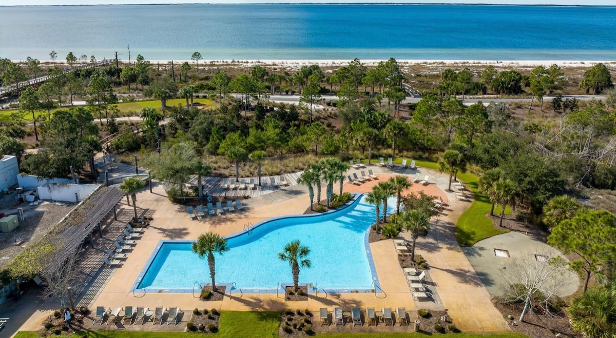 A resort-style community pool is surrounded by lush greenery and palm trees, with numerous lounge chairs placed along the water's edge. The pool's clear blue water reflects a sunny sky, offering a refreshing contrast to the sandy beach visible in the background.