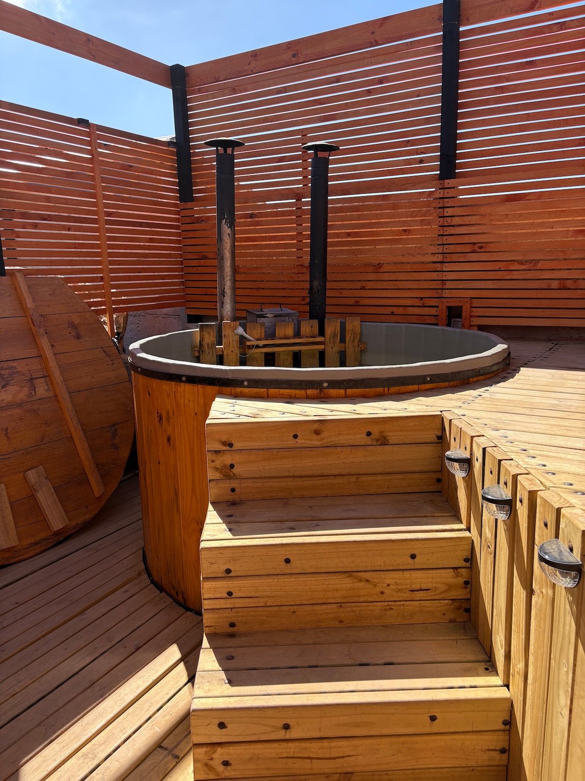 A wooden hot tub is positioned on a deck surrounded by slatted wooden fencing, providing privacy. The tub is accessed via wooden steps. Additional wooden elements are visible, creating a natural and rustic ambiance in the outdoor space.