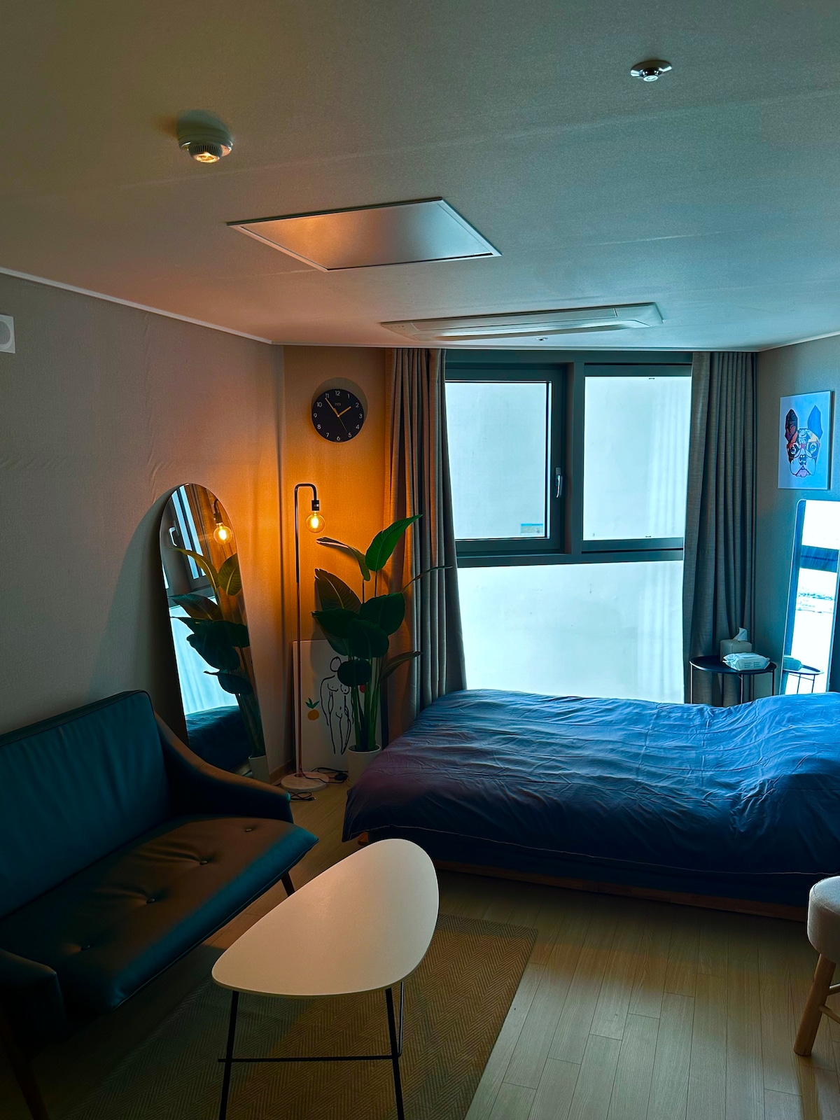 A cozy bedroom with a spacious bed is complemented by soft blue bedding. A mirror is positioned beside the bed, reflecting ambient lighting from a floor lamp. A small table and a dark sofa introduce a relaxing seating area, while a window offers natural light.