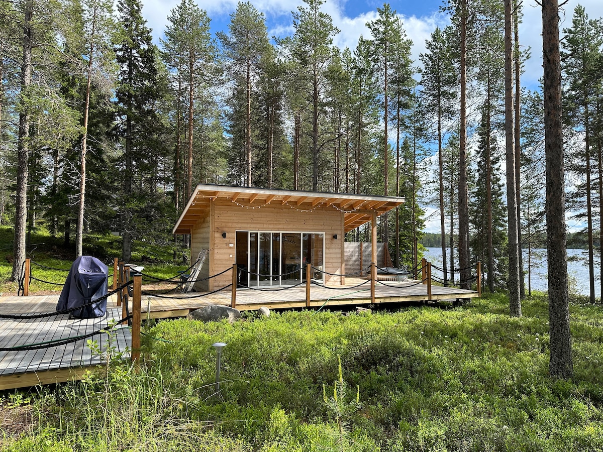 A modern cabin is surrounded by tall pine trees, featuring large glass doors that open to a wooden deck. The deck is equipped with a hammock and has a serene view of the lake just beyond the trees, creating a peaceful outdoor space.