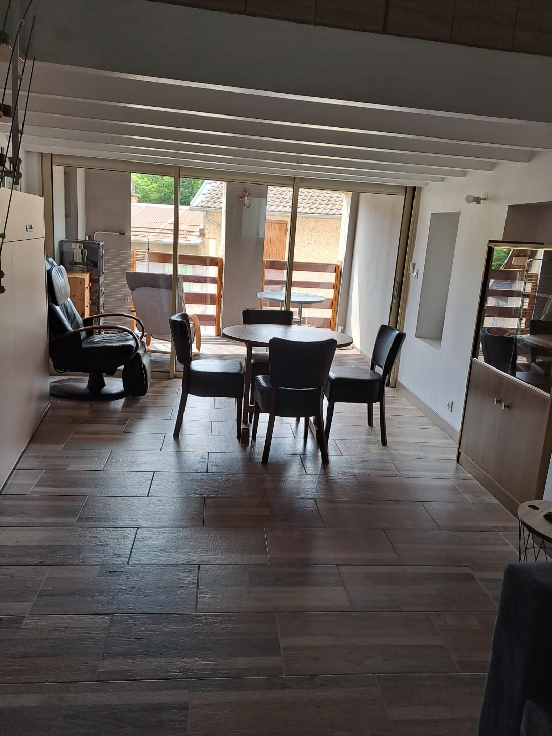 A bright dining area features a round table surrounded by four chairs. Large glass doors provide natural light and open to a balcony with views of the outdoors. A comfortable chair and a side cabinet are also visible, enhancing the spaciousness.
