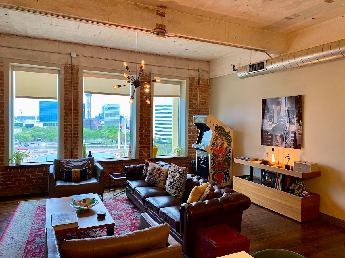 A relaxing living area features a brown leather sectional sofa and an armchair, arranged around a coffee table. Large windows offer views of the cityscape, while an eclectic chandelier adds character. A vintage arcade game and a console occupy opposite corners, enhancing the inviting space.