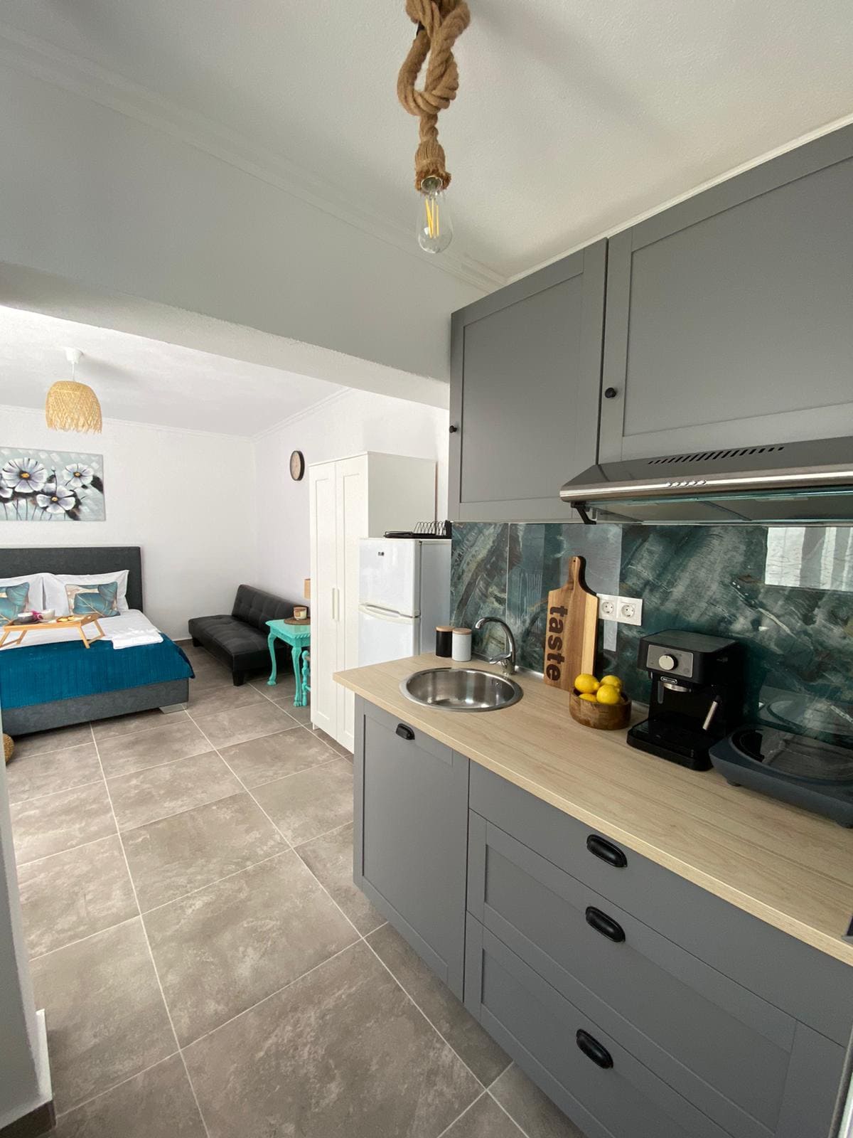 A modern kitchenette is shown, featuring gray cabinetry and a wooden countertop. A small sink and kitchen appliances are positioned alongside a dining area. A cozy sleeping area with a bed and seating is visible in the background, offering a clean and contemporary atmosphere.
