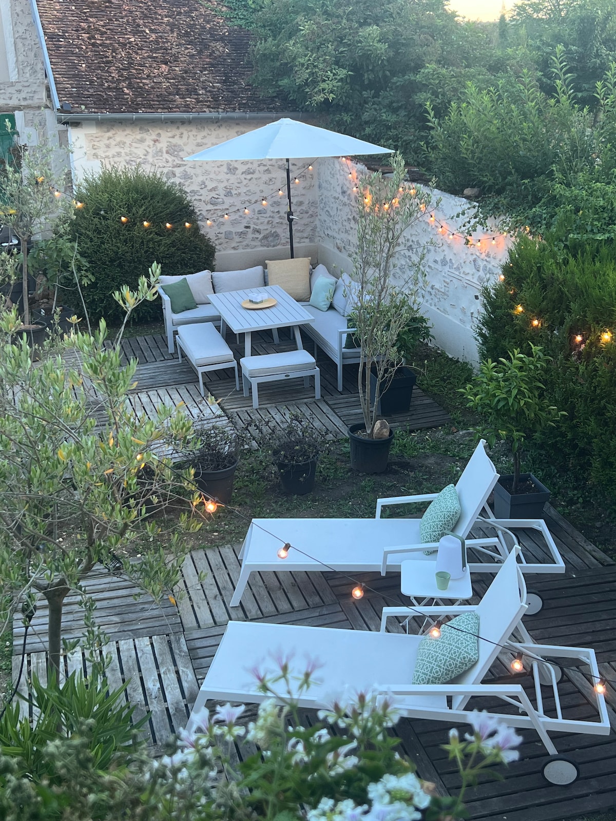A serene garden space is presented, featuring a wooden deck surrounded by greenery. Cozy seating is arranged with a parasol overhead. Soft lighting from string lights enhances the ambiance. Lounge chairs invite relaxation in a tranquil outdoor setting.