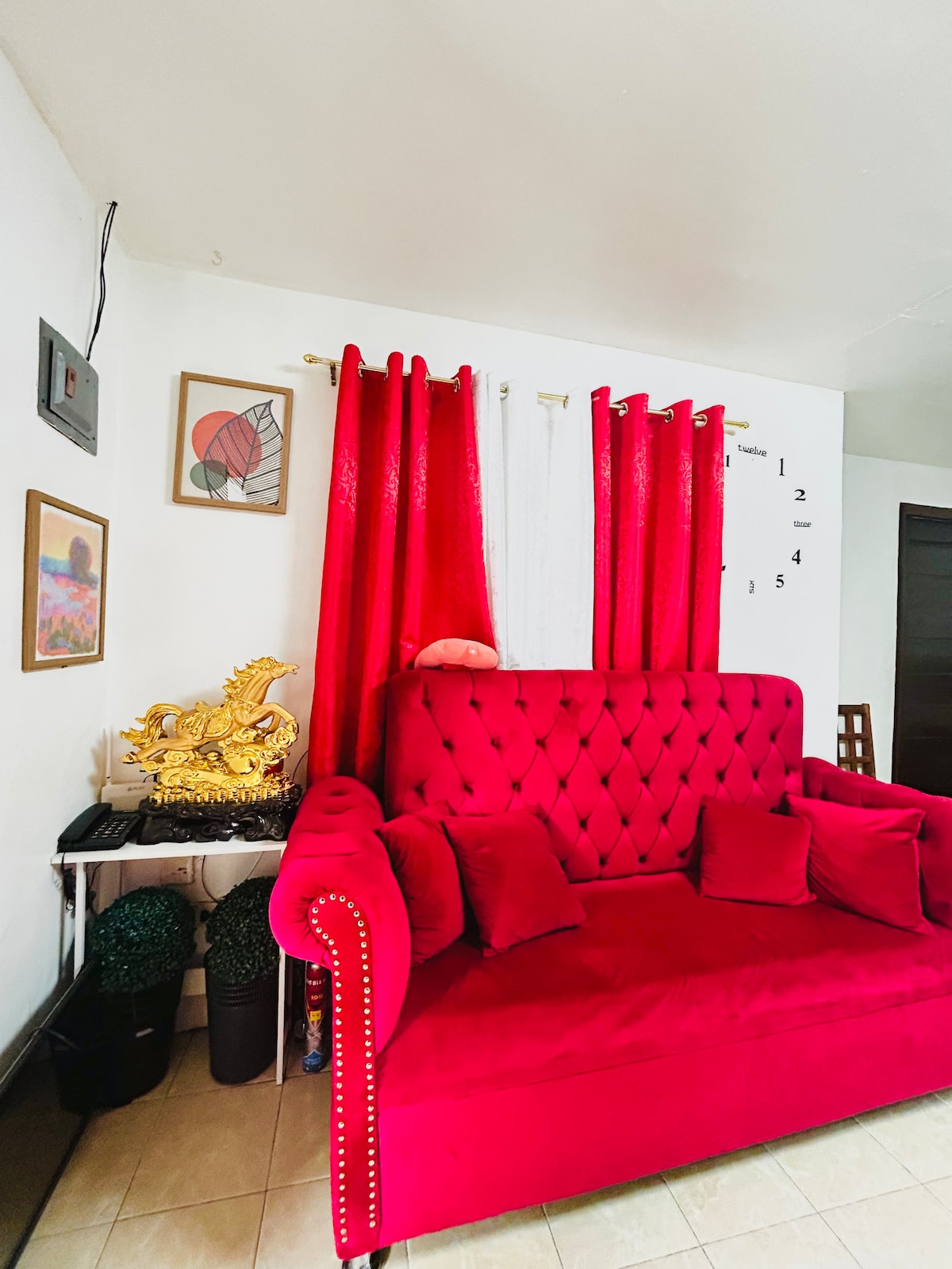 A cozy living space features a vibrant red tufted sofa, accented with several matching cushions. Light filters through sheer and curtain panels, complementing the wall decor, which includes framed artwork. A decorative golden figure rests on a nearby surface amidst potted greenery.