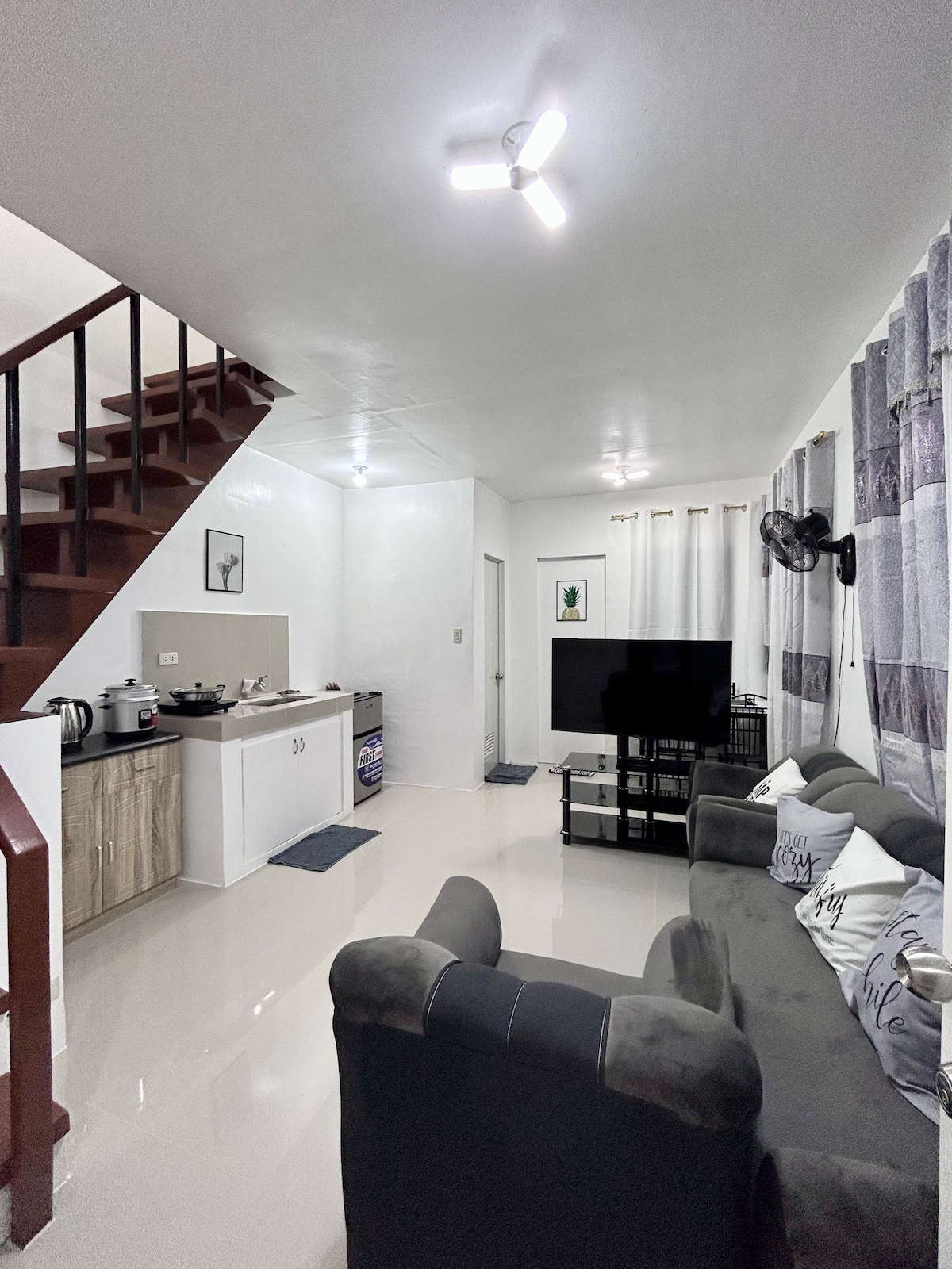 An open living area is captured, featuring a comfortable sofa facing a black TV stand with a large screen. A staircase ascends to the upper level, while a well-equipped kitchenette is positioned in the back, illuminated by soft overhead lighting.