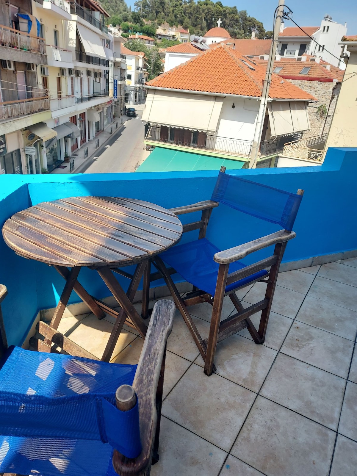 A balcony space is highlighted by a round wooden table and two blue folding chairs. The tiled floor complements the vibrant blue walls. A view of neighboring buildings and a sunny street is visible, creating a pleasant outdoor setting.