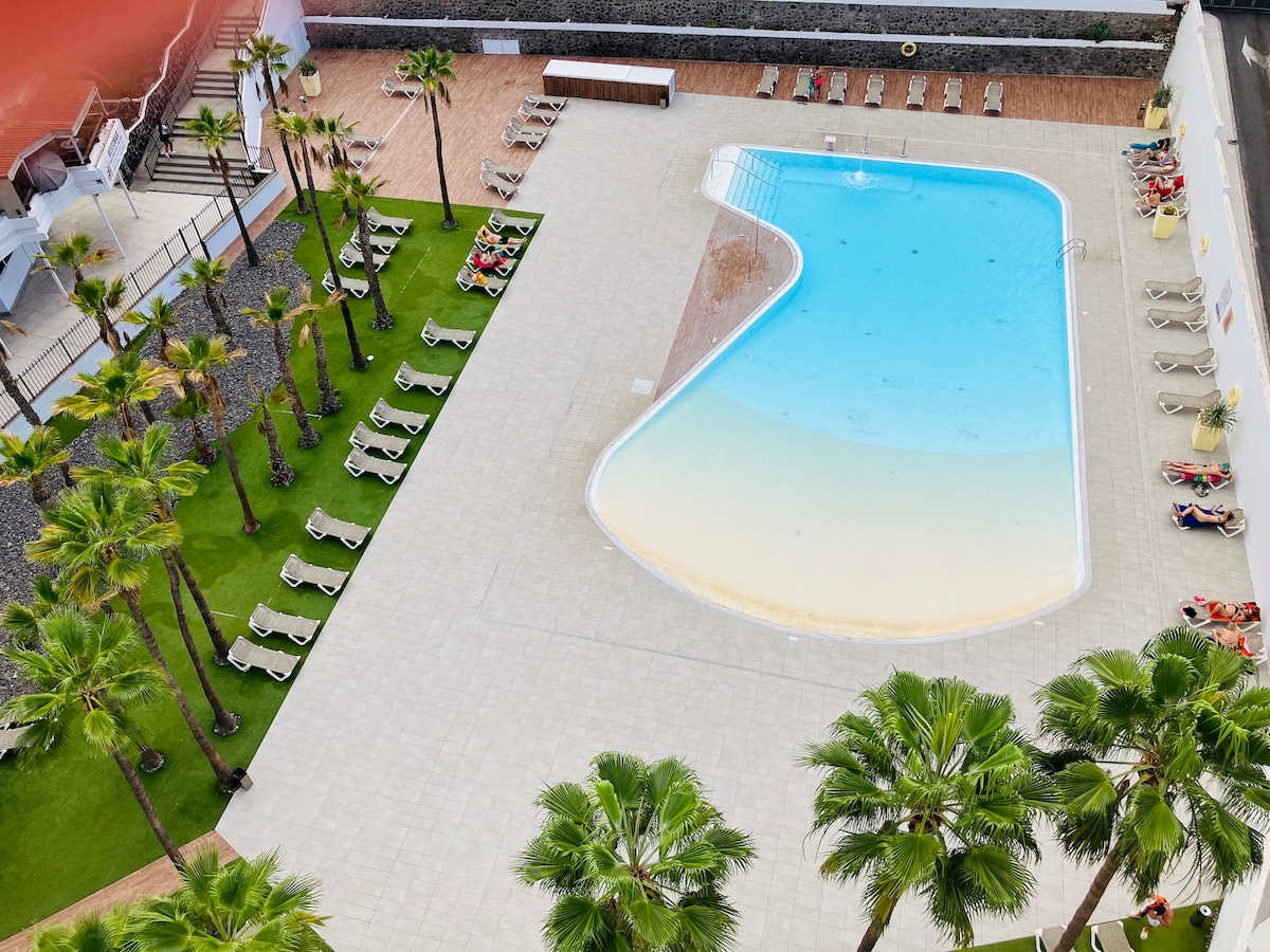 A spacious outdoor area features a large swimming pool with a gradual entry. Surrounding palms provide shaded spots, while several lounge chairs are arranged neatly along the poolside. The area includes well-maintained green grass, enhancing the inviting ambiance.