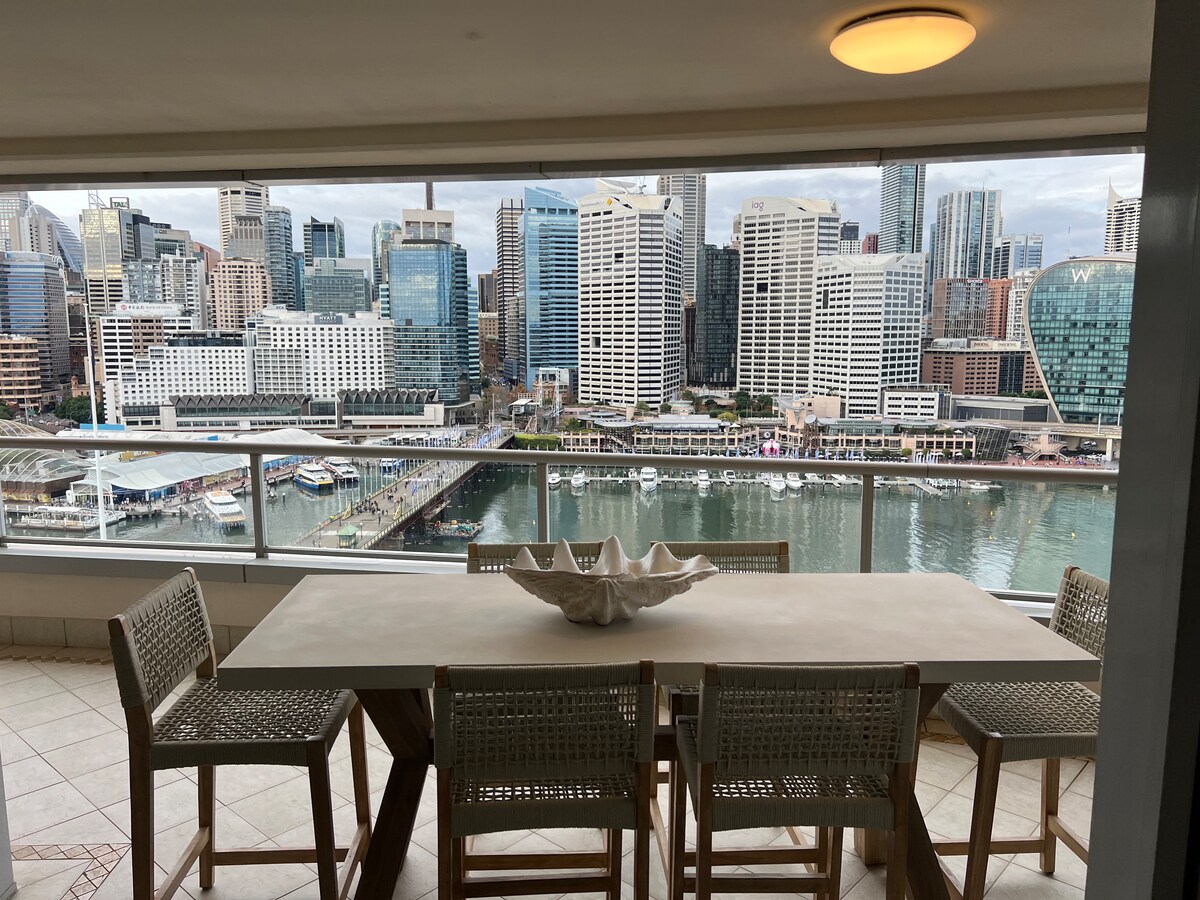A spacious balcony dining area features a large table accompanied by six woven high-back chairs. The panoramic view showcases the bustling Darling Harbour, surrounded by modern skyscrapers reflecting in the water, creating a vibrant cityscape.
