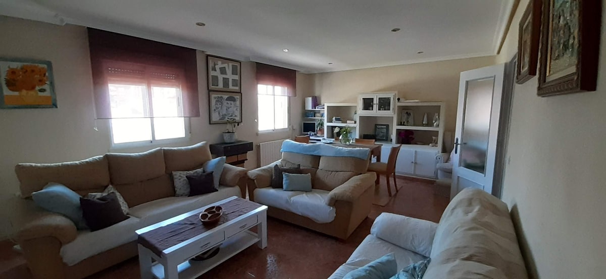 The spacious living area is furnished with two light-colored sofas and a white coffee table at the center. A dining table with chairs is visible in the background. Large windows allow natural light to fill the space, creating a bright and airy atmosphere.
