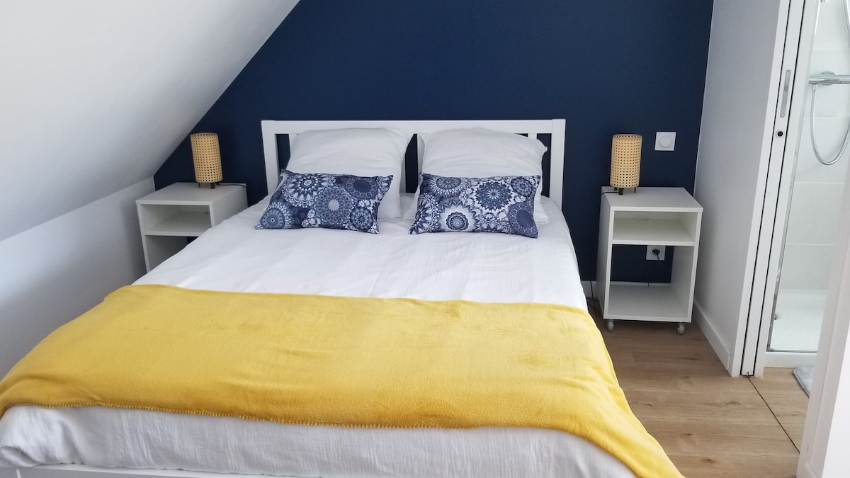 A bright bedroom features a spacious bed adorned with soft white linens and a yellow throw blanket. Decorative pillows add a touch of color, while two bedside tables with lamps provide illumination. A glass-enclosed shower is visible in the adjacent area.