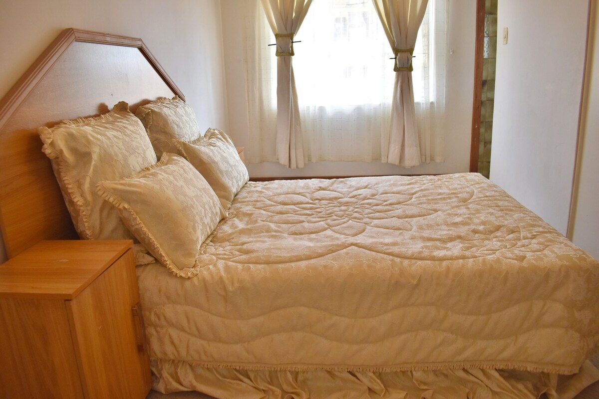 A spacious bed is adorned with a light-colored quilt featuring a floral pattern. Soft, decorative pillows rest against the headboard. Natural light filters through sheer curtains, illuminating the room, which includes a wooden nightstand on one side.
