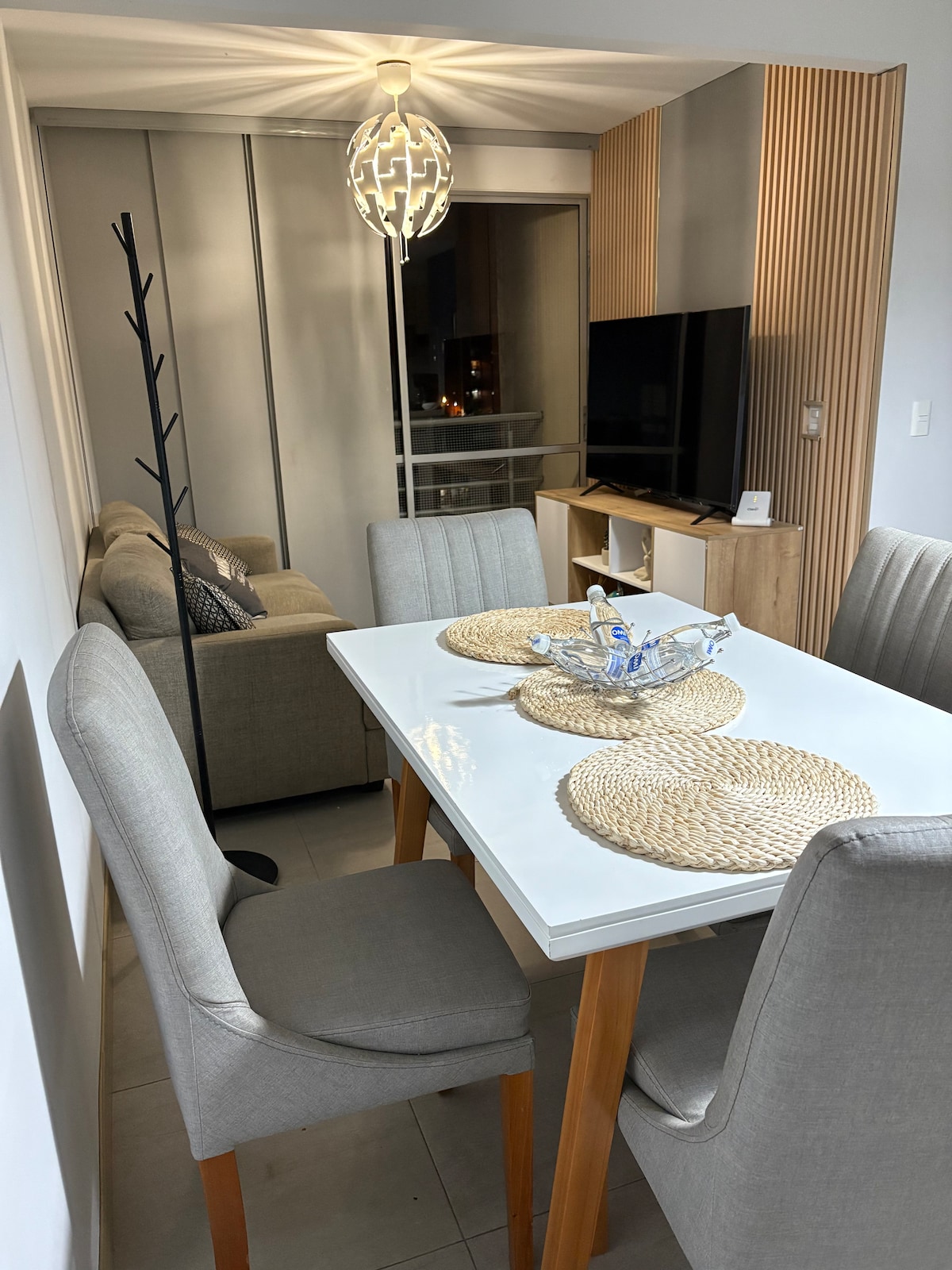 A modern dining area features a white table surrounded by four upholstered chairs. A decorative bowl sits at the center of the table, complemented by woven placemats. In the background, a stylish sofa overlooks a television, with warm lighting enhancing the space.