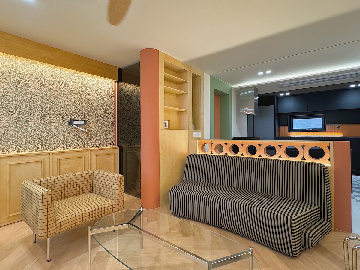 A modern living room features a striped sofa and a patterned armchair on a light wood floor. Warm light illuminates the walls, showcasing a decorative panel. A glass coffee table sits in front, complementing the contemporary decor and layout of the space.