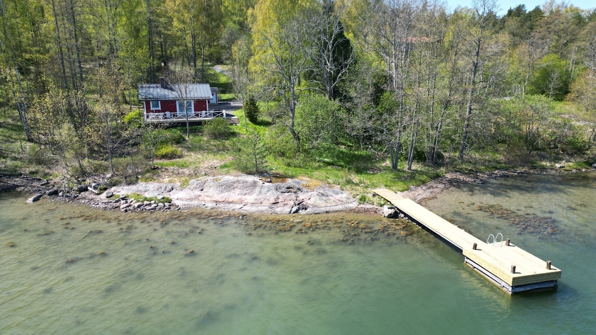 A serene lakeside setting showcases a cozy red cabin nestled among lush greenery. A wooden dock extends into the water, providing access for swimming. Rocky shorelines blend naturally with calm waters, framed by trees that enhance the peaceful atmosphere of the area.