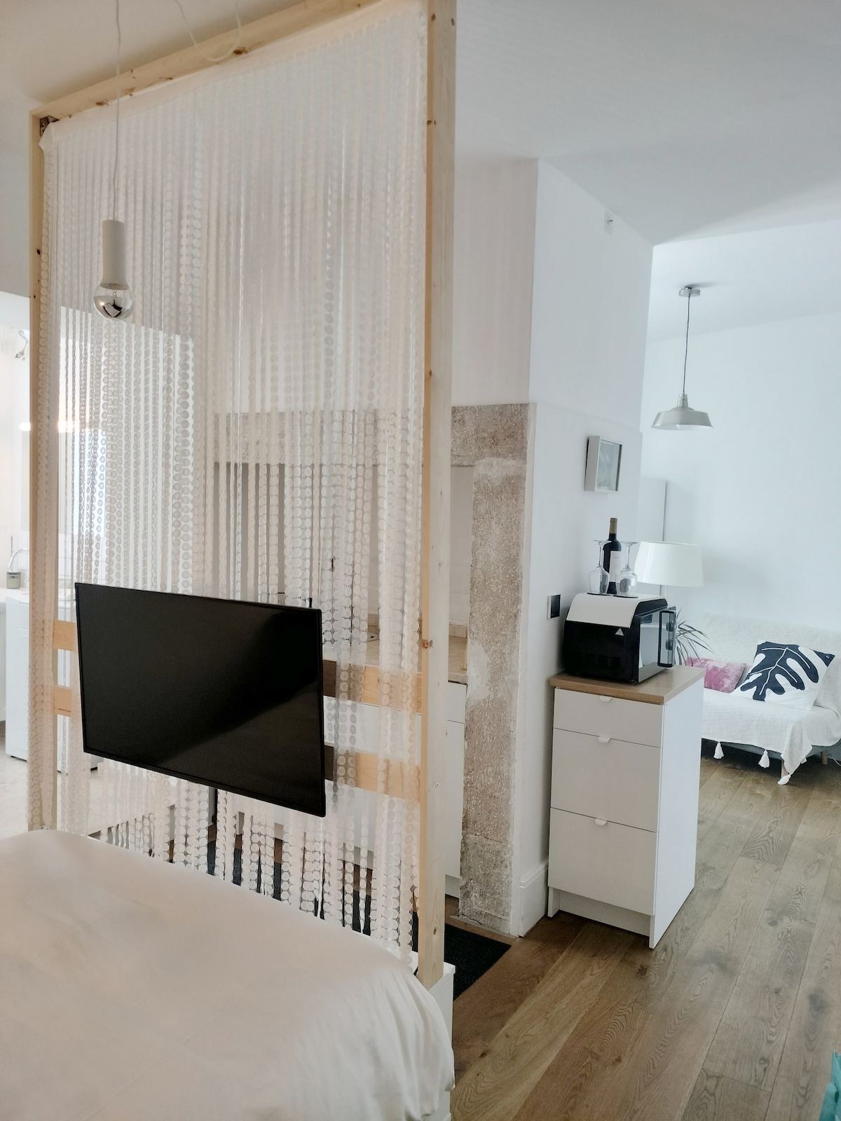 A space divider made of hanging beads is positioned near a wall-mounted television, creating a sense of separation between areas. A kitchenette is visible to the left, while a light-colored sofa is seen in the background. Natural light floods the room, highlighting the wooden flooring.