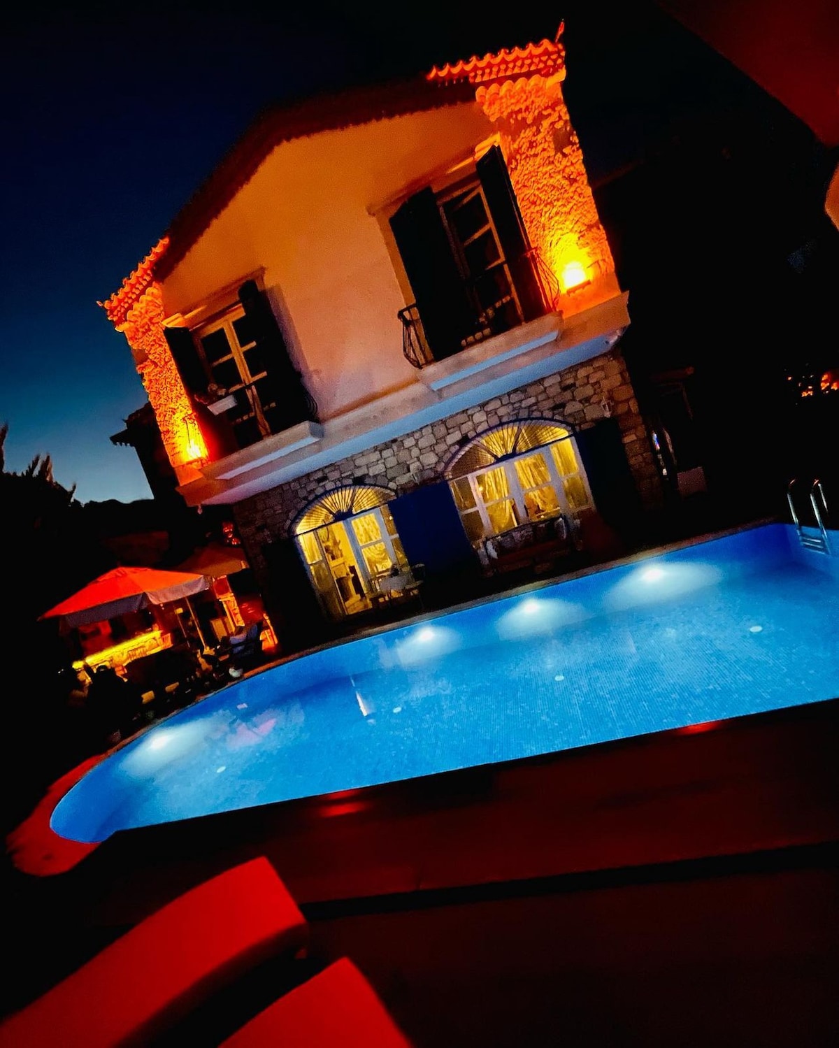An illuminated pool area is complemented by a stone building featuring large glass windows. The turquoise water of the pool glistens under soft lighting, while surrounding furniture and umbrellas create a welcoming outdoor space for relaxation.